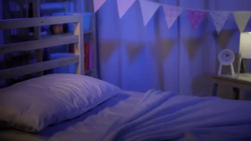غرفة نوم مضاءة بخافت، تحتوي على سرير مع وسادة وغطاء، وزينة ملونة معلقة، تعكس حالة القلق التي عاشها الطفل.