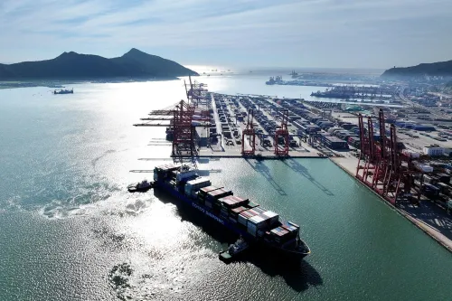 صورة جوية لميناء مزدحم، تُظهر سفينة شحن محملة بالحاويات، مع رافعات ومرافق تخزين في الخلفية، تعكس أهمية التجارة بين الصين والولايات المتحدة.