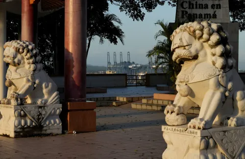 تمثالان من الحجر الأبيض لأسود صينية تقفان في حديقة في بنما، مع خلفية تظهر موانئ ومعدات شحن، مما يعكس تأثير الصين في أمريكا اللاتينية.