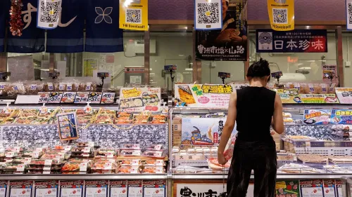 امرأة تتسوق في متجر للمأكولات البحرية، معروضات متنوعة من الأطعمة البحرية اليابانية، تعكس تأثيرات قرار الصين بشأن الاستيراد.