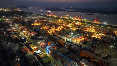 مشهد ليلي لميناء مزدحم بالحاويات، حيث تُظهر الأضواء والشاحنات الحركة النشطة في ظل التصعيد التجاري بين الولايات المتحدة والصين.
