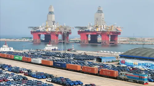 صورة لميناء مزدحم يظهر فيه سفن ضخمة للتنقيب عن النفط، مع شاحنات وقطارات تحمل حاويات، ومركبات متوقفة في الخلفية.