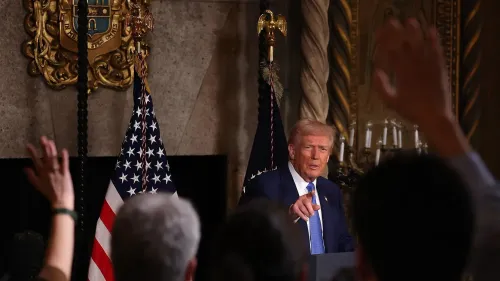 ترامب يتحدث في مؤتمر صحفي، مع رفع الأيدي من الحضور، وعلم الولايات المتحدة خلفه، يعكس تفاعلاته مع الجمهور حول قضايا سياسية.