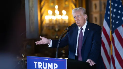 دونالد ترامب يتحدث في مؤتمر صحفي، مع لافتة تحمل اسمه خلفه، وعلم أمريكي يظهر إلى جانبه، في سياق استعداداته لفترة ولايته الثانية.