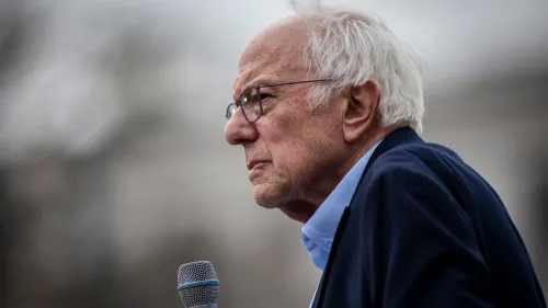 السيناتور بيرني ساندرز يتحدث في حدث عام، مع ميكروفون في يده، وسط خلفية ضبابية تعكس أجواء السياسة الأمريكية.