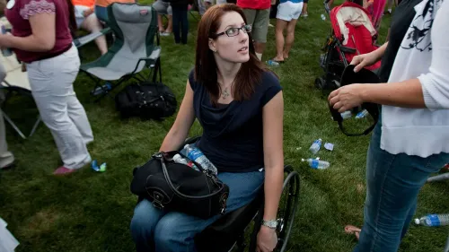 امرأة جالسة في كرسي متحرك، تنظر حولها في تجمع خارجي، تعكس قوتها وإيجابيتها رغم تحدياتها الصحية بعد حادث إطلاق النار في كولومباين.