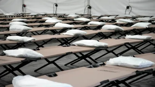 صفوف من أسرة مؤقتة مزودة بوسائد داخل خيمة، تعكس جهود توفير المأوى للمهاجرين في ظل مناقشات حول إعادة تخصيص التمويل.