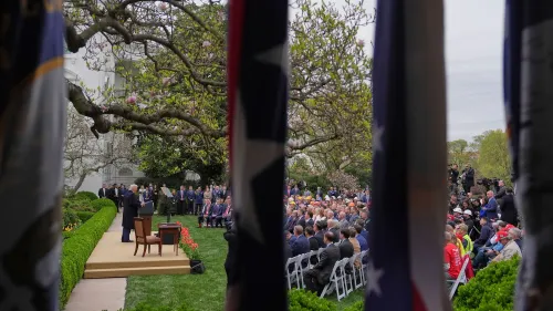 اجتماع في حديقة البيت الأبيض حيث يتحدث الرئيس ترامب أمام حشد من الحضور، مع وجود أعلام في الخلفية وأشجار مزهرة.