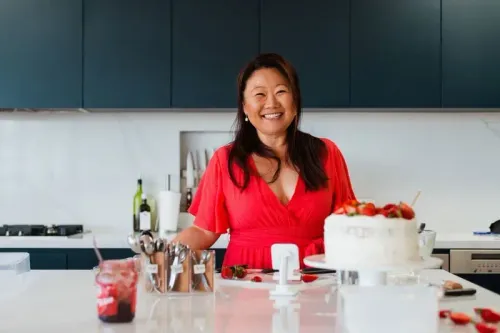 امرأة مبتسمة ترتدي فستاناً أحمر وتقف في مطبخ حديث بجوار كعكة مزينة بالفراولة، تعكس شغفها بالخبز والإبداع.