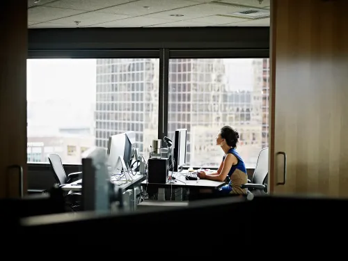 امرأة تعمل على جهاز كمبيوتر في مكتب حديث، مع إطلالة على ناطحات السحاب من النافذة، تعكس تحديات النساء في المناصب القيادية.