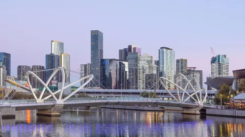 منظر لمدينة ملبورن مع نهر وجسر حديث، يظهر العمارة العصرية والمباني الشاهقة في خلفية الغروب.