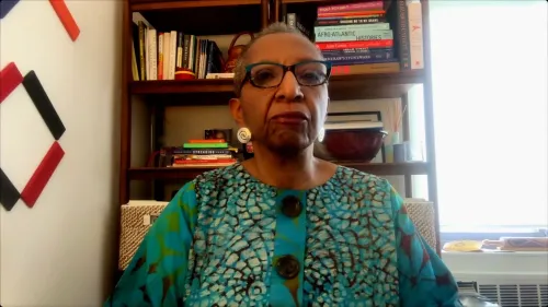 امرأة ترتدي قميصًا مزخرفًا باللونين الأزرق والأخضر، تجلس أمام رف مليء بالكتب، تعبر عن قضايا قانونية تتعلق بفصل المسؤولين الفيدراليين.