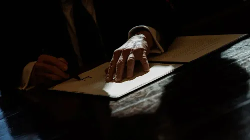 يد تظهر يد شخص تكتب على ورقة، مع تسليط الضوء على الأثر السياسي والاقتصادي للقرارات الحكومية.