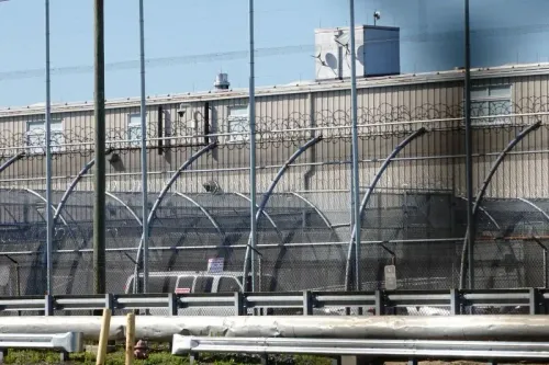 سياج حديدي يحيط بمركز احتجاز للمهاجرين، يظهر المبنى الرئيسي مع أسلاك شائكة، مما يعكس الظروف القاسية للقبض على المهاجرين.