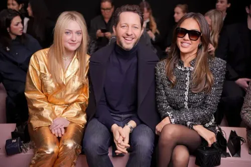 ثلاثة أشخاص يجلسون في الصف الأمامي خلال عرض أزياء في باريس، يرتدي أحدهم بدلة ذهبية، بينما الأخرى ترتدي ملابس سوداء أنيقة، مع ابتسامات واضحة.