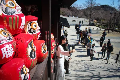 مجموعة من دمى داروما الحمراء في معبد كاتسو-جي، مع زائر يأخذ صورة سيلفي، تعكس رمزية الحظ والطموح في الثقافة اليابانية.