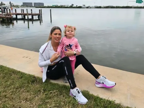 هايدي سانشيز تجلس على ضفة مائية مع ابنتها الصغيرة، مرتدية ملابس رياضية، وتبتسم بينما تلتقطان لحظة عائلية مع خلفية طبيعية.