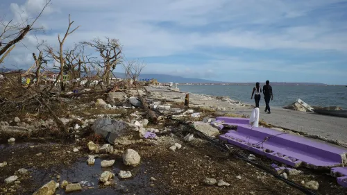 تظهر الصورة مشهدًا مدمّرًا في جامايكا بعد إعصار ميليسا، مع بقايا المنازل والأشجار المكسورة، بينما يسير شخصان على الواجهة البحرية.