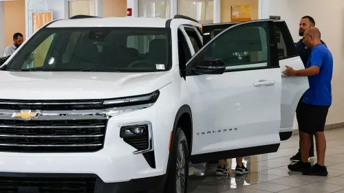شخصان يتفقدان سيارة شيفروليه ترافيرس بيضاء في صالة عرض للسيارات، مع التركيز على تفاصيل السيارة الحديثة في ظل توقعات ارتفاع الأسعار.