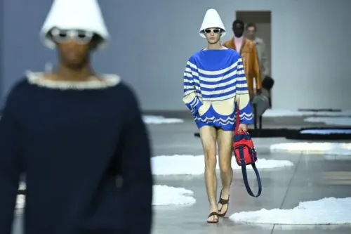 عارض أزياء يرتدي سترة زرقاء مخططة وشورت قصير، يسير على منصة عرض الأزياء في أسبوع الموضة بميلانو، مع خلفية فنية.