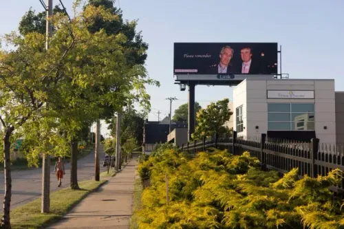 لافتة إعلانات خارجية تظهر صورة لجيفري إبشتاين ودونالد ترامب مع عبارة "يرجى التذكر"، وسط أشجار وأماكن عامة.