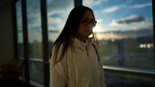 امرأة ترتدي سترة بيضاء، واقفة بجانب نافذة كبيرة، مع غروب الشمس في الخلفية، تعبر عن مشاعر القلق بشأن مرض الذئبة.