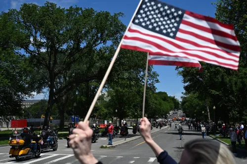 رجل يحمل علم الولايات المتحدة خلال مسيرة لدعم قدامى المحاربين، مع وجود أشخاص ومركبات في الخلفية، تعبيرًا عن التضامن والمساندة.