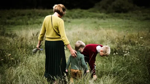 امرأة ترتدي سترة صفراء وسروال طويل أسود، ترافق طفلين في حقل أخضر، حيث ينشغل أحدهما بجمع الأعشاب.