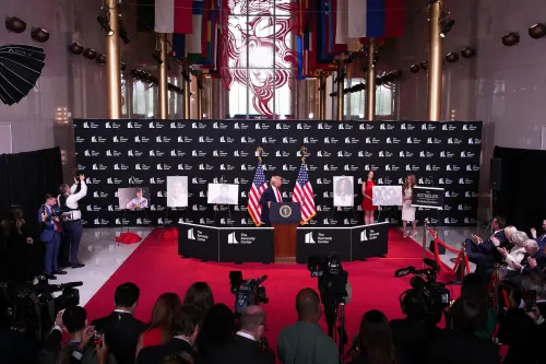 ترامب يتحدث في مركز كينيدي، محاطًا بالعلم الأمريكي وصور المكرمين، بينما يلتقط الصحفيون الصور.