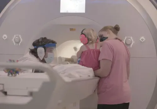 طفلة صغيرة ترتدي سماعات أذن، تُحتضَر من قبل والدتها أثناء إجراء فحص بالرنين المغناطيسي، مع وجود باحث بجانبهم.