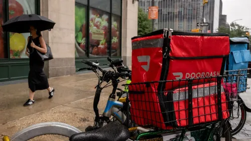 امرأة تحمل مظلة تسير بجانب دراجة مزودة بحقيبة توصيل حمراء تحمل شعار DoorDash، في مشهد يعكس خدمات توصيل الوجبات في المدينة.