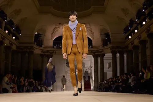 رجل يرتدي بدلة بنية وحذاء بكعب عالٍ، يسير على منصة عرض أزياء في باريس، مع جمهور ومصممين في الخلفية.