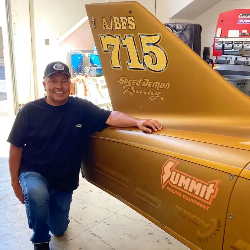 سائق سباق مبتسم يجلس بجانب سيارة "Speed Demon" ذات اللون الذهبي، مع تفاصيل واضحة للعلامات التجارية والمعدات المستخدمة في السباق.