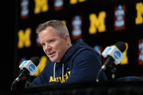 مدرب فريق ولفيرينز، داستي ماي، يتحدث في مؤتمر صحفي، مع خلفية تحمل شعار جامعة ميشيغان، مع التركيز على نجاح الفريق في كرة السلة الجامعية.