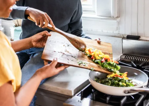 تحضير وجبة صحية في المطبخ، حيث يقوم شخصان بإضافة الخضروات الطازجة إلى مقلاة، مما يعكس أهمية الطعام المعالج بالحد الأدنى.