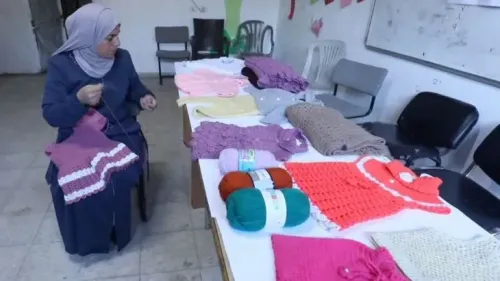 امرأة فلسطينية تعمل على حياكة الملابس في غرفة بسيطة، محاطة بأقمشة ملونة ومنتجات جاهزة، تعكس جهود البقاء وسط الأزمة الاقتصادية في غزة.