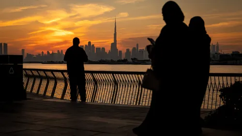غروب الشمس فوق أفق دبي، مع silhouettes لأشخاص على ضفاف الماء، وتظهر برج خليفة في الخلفية، مما يعكس جمال المدينة.