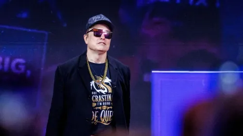 إيلون ماسك يرتدي نظارات شمسية وقميصًا مكتوبًا عليه "لا أؤجل"، مع خلفية ملونة، أثناء حديثه في حدث عام.