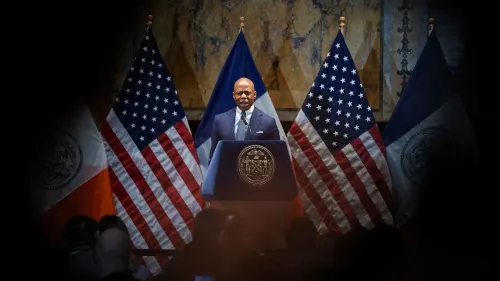 إريك آدامز يتحدث أمام منصة تحمل شعار مدينة نيويورك، محاطًا بأعلام الولايات المتحدة ومدينة نيويورك، في ظل تصاعد الضغوط المطالبة باستقالته.