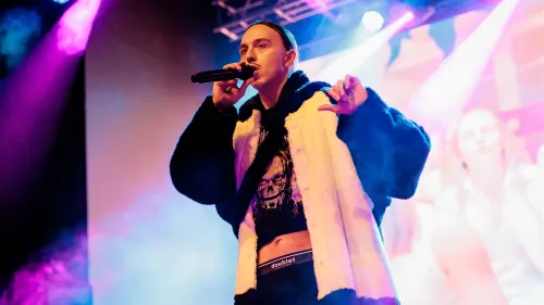 مغني الراب تومي كاش يؤدي على المسرح، مرتديًا معطفًا أسود، ويستخدم ميكروفونًا، وسط أضواء ملونة، في سياق جدل حول أغنيته "إسبريسو ماتشياتو".