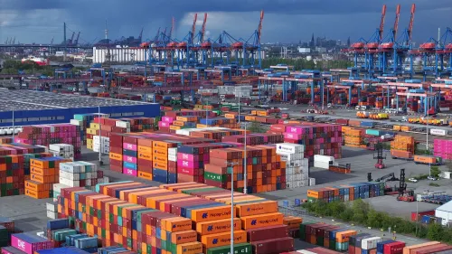 مستودع شحن مزدحم يحتوي على حاويات ملونة، مع رافعات تعمل في الخلفية، يعكس تأثير الحرب التجارية على الاقتصاد الأوروبي.