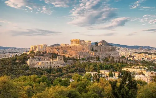 أكروبوليس أثينا يبرز فوق المدينة، محاطًا بالأشجار، ويظهر خلفه مشهد حضري مع السماء الزرقاء. يعكس الصورة تحديات الحرارة الصيفية.