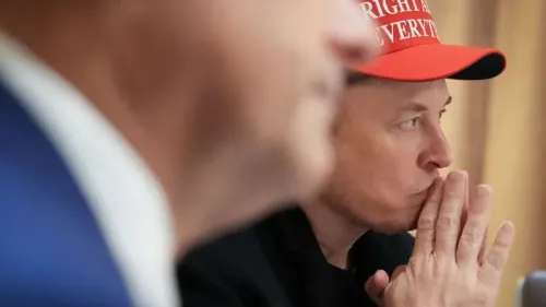 إيلون ماسك يفكر بجدية خلال اجتماع، مرتديًا قبعة تحمل شعارًا سياسيًا، مما يعكس تأثيره المتزايد على علامة تسلا التجارية.