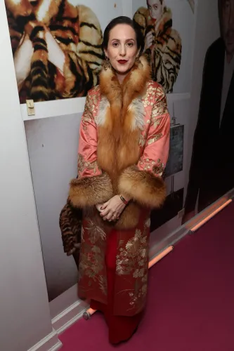 امرأة ترتدي معطفًا مزخرفًا بالفرو، تقف أمام جدار مزين بصور، تعكس عودة الفرو إلى الموضة مع التركيز على الاستدامة.