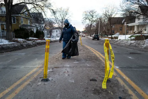 شخص يجمع القمامة في شارع مغلق بشريط شرطة أصفر، مع بقايا ثلوج وأشجار في الخلفية، مما يعكس جهود التنظيف في منطقة متأثرة بالاضطرابات.