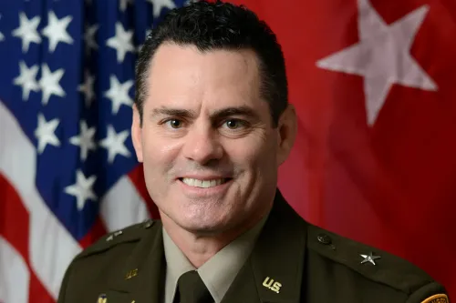 صورة للعميد إريك ويدمار، محامي هيئة الأركان المشتركة، مبتسمًا أمام علم الولايات المتحدة، مع خلفية حمراء ونجوم بيضاء.