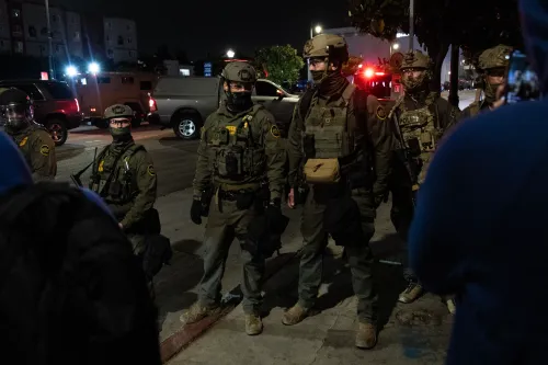 مجموعة من عناصر إنفاذ القانون يرتدون زيًا عسكريًا، يتواجدون في موقع اعتقال ليلي، مع وجود سيارات في الخلفية.