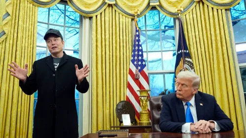 إيلون ماسك يتحدث في المكتب البيضاوي بينما يجلس دونالد ترامب، مع خلفية من الستائر الذهبية وعلمين أمريكيين.