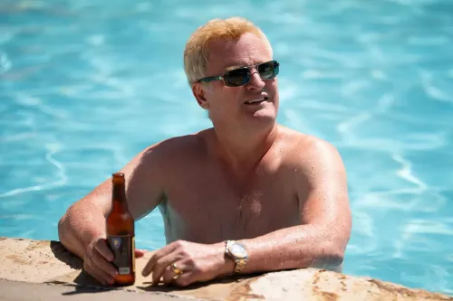 رجل عائم في بركة سباحة، يرتدي نظارات شمسية، ممسكًا بزجاجة مشروب، في مشهد يعكس حياة الرفاهية قبل الأحداث المأساوية لعائلة موردو.