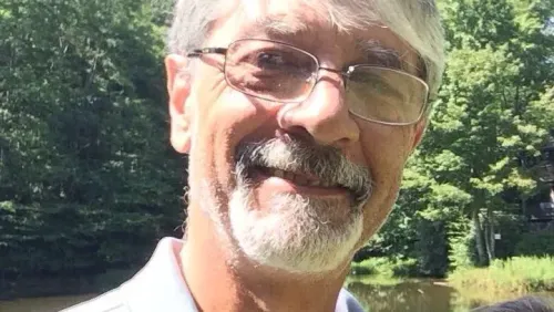 صورة لرجل مسن ذو لحية وشعر رمادي، يرتدي نظارات، مبتسم في بيئة طبيعية. تعكس الصورة معاناة محتجزين في مركز الهجرة في فلوريدا.